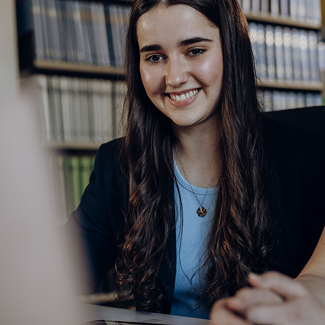 Junge Frau sitzt vor Computer in einer Bücherei und lächelt